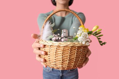 Elinde hasır Paskalya sepeti ve pembe arka planda kozmetik ürünleri tutan genç bir kadın.