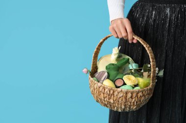 Mavi arka planda kozmetik ürünleri olan hasır Paskalya sepeti tutan genç bir kadın.