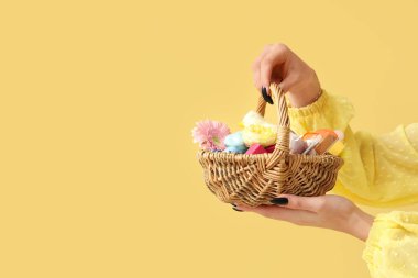 Sarı arka planda kozmetik ürünleri olan hasır bir Paskalya sepeti tutan genç bir kadın.
