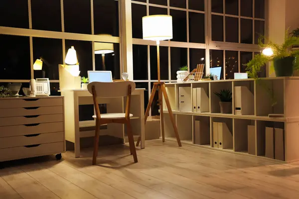 Interior of office with workplace, shelf unit and glowing lamps at night