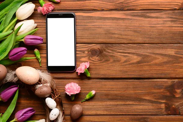 Modern cep telefonlu kompozisyon, güzel çiçekler ve tahta arka planda Paskalya çelengi.