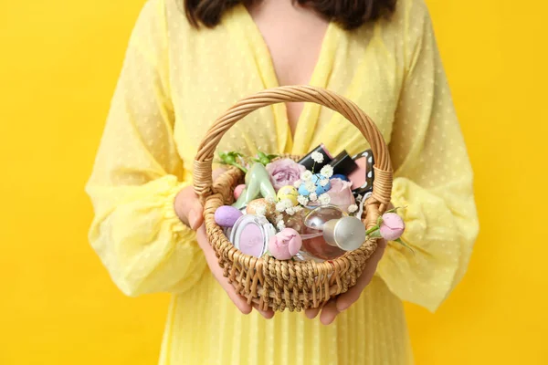 Woman holding Easter basket with cosmetic products on yellow background, closeup