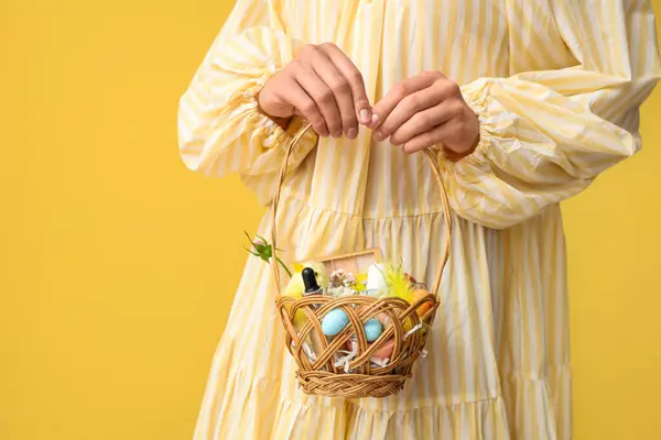 Sarı arka planda kozmetik ürünleri olan hasır bir Paskalya sepeti tutan genç bir kadın.