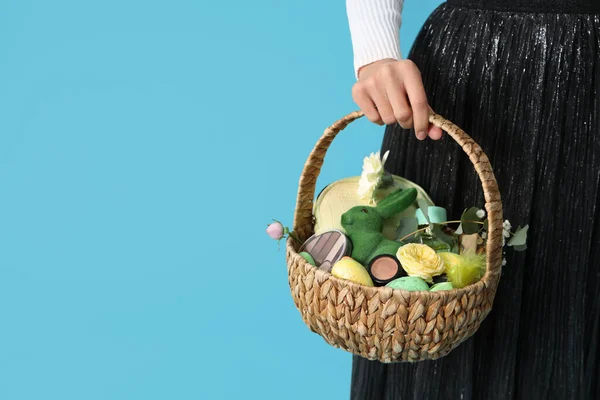 Mavi arka planda kozmetik ürünleri olan hasır Paskalya sepeti tutan genç bir kadın.