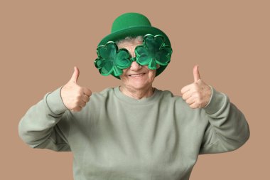 Leprikon şapkalı ve yonca şeklinde dekoratif gözlüklü yaşlı kadın kahverengi arka planda baş parmak hareketi gösteriyor. Aziz Patrick Günü kutlaması