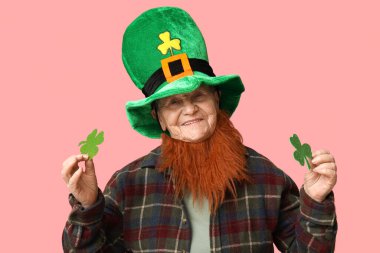Kırmızı sakallı cüce şapkalı yaşlı kadın pembe arka planda yonca tutuyor. Aziz Patrick Günü kutlaması