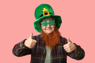 Kırmızı sakallı, cüce şapkalı ve dekoratif gözlüklü yaşlı kadın pembe arka planda baş parmak hareketi gösteriyor. Aziz Patrick Günü kutlaması