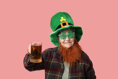 Leprikon şapkalı, dekoratif gözlüklü, kırmızı sakallı pembe arka planda bir bardak bira tutan yaşlı kadın. Aziz Patrick Günü kutlaması