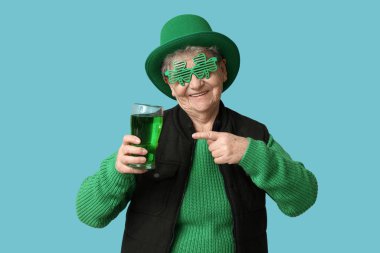Leprikon şapkalı ve dekoratif gözlüklü yaşlı kadın mavi arka planda bir bardak biraya bakıyor. Aziz Patrick Günü kutlaması