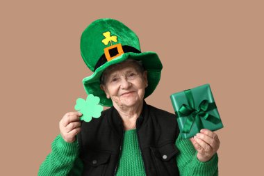 Kahverengi arka planda yonca ve hediye kutusu olan cüce şapkalı son sınıf kadın. Aziz Patrick Günü kutlaması