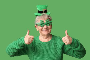 Leprikon şapkalı ve dekoratif gözlüklü yaşlı kadın yeşil arka planda baş parmak hareketi gösteriyor. Aziz Patrick Günü kutlaması