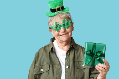 Leprikon şapkalı, dekoratif gözlüklü, mavi arka planda hediye kutusu olan yaşlı bir kadın. Aziz Patrick Günü kutlaması