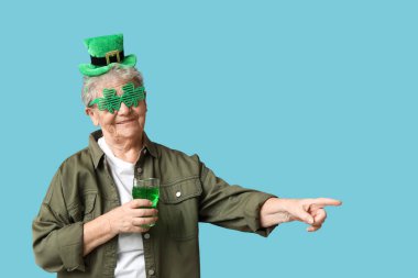 Leprikon şapkalı ve dekoratif gözlüklü yaşlı kadın mavi arka plandaki bir şeyi işaret eden bir bardak birayla. Aziz Patrick Günü kutlaması