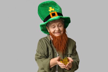 Kırmızı sakallı cüce şapkalı yaşlı kadın gri arka planda bozuk para tutuyor. Aziz Patrick Günü kutlaması