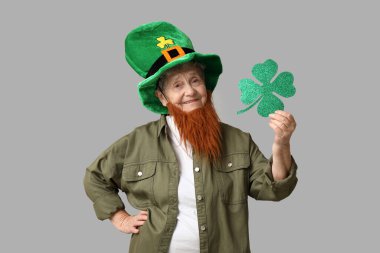 Kırmızı sakallı cüce şapkalı yaşlı kadın gri arka planda yonca tutuyor. Aziz Patrick Günü kutlaması