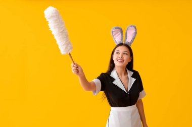 Tavşan kulaklı genç Asyalı oda hizmetçisi ve sarı arka planda toz bezi. Paskalya kutlaması