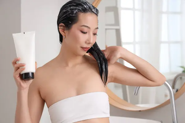 Young Asian woman with tube of hair mask in bathroom, closeup