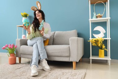 Tavşan kulaklı genç bir kadın. Paskalya tavşanı ve yumurtalı. Evdeki kanepede oturuyor.