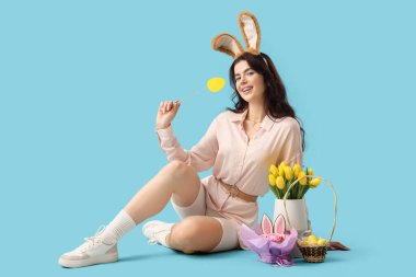 Tavşan kulaklı güzel bir kadın. Paskalya yumurtası, pasta ve laleler mavi arka planda oturuyor.