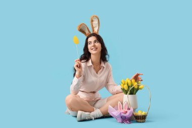 Tavşan kulaklı güzel bir kadın. Paskalya yumurtası, pasta ve laleler mavi arka planda oturuyor.