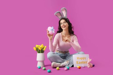Paskalya yumurtaları ve laleleri mor arka planda oturan tavşan kulaklı güzel bir kadın.