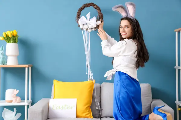 Tavşan kulaklı genç kadın evdeki mavi duvara Paskalya çelengi asıyor.