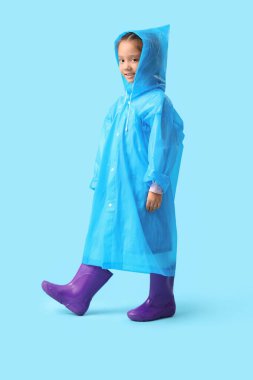 Yağmurluklu küçük Asyalı kız ve mavi arka planda lastik çizmeler.