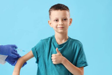 Küçük çocuk aşı oluyor ve mavi arka planda baş parmağını kaldırıyor.