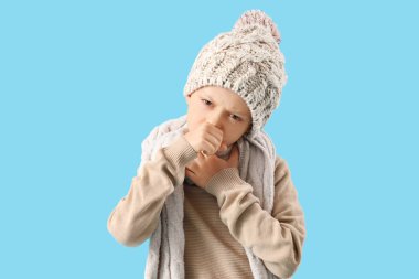Mavi arka planda şapkalı küçük çocuk öksürüyor.