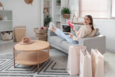 Hediye çeki ve dizüstü bilgisayarıyla evdeki kanepede alışveriş yapan mutlu genç bir kadın.