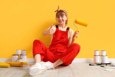 Boya silindirli kadın dekoratör odada tamirat sırasında başparmağını kaldırıyor.