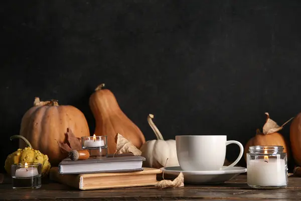 Cup of tea, books, autumn leaves, burning candles and pumpkins on table near black wall