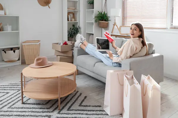Hediye çeki ve dizüstü bilgisayarıyla evdeki kanepede alışveriş yapan mutlu genç bir kadın.