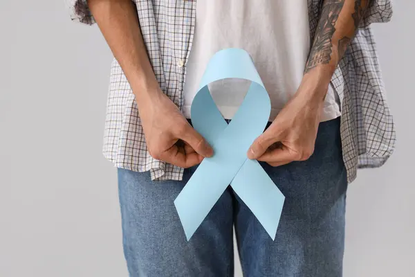 Young man with blue ribbon on light background, closeup. Prostate cancer awareness concept