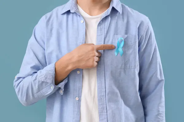 Young man pointing at blue ribbon on color background, closeup. Prostate cancer awareness concept