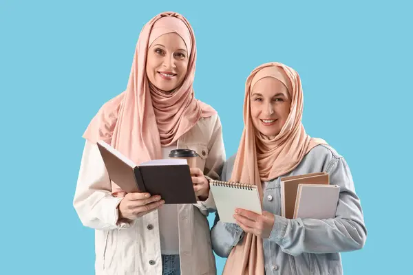 Mature Muslim women in hijabs with books and coffee on blue background