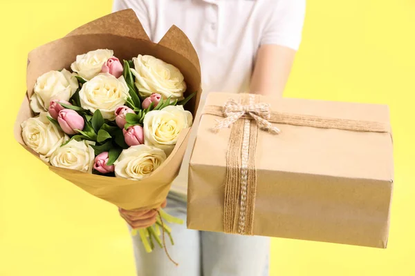 Female courier with bouquet of flowers and gift box on yellow background, closeup