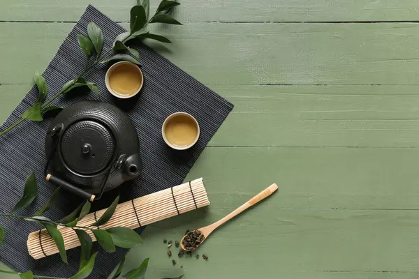 Çay fincanlı bambu hasırı, çaydanlık ve yeşil arka planda kuru yapraklı kaşık.