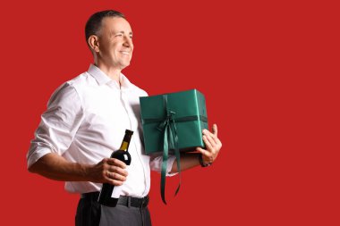 Kırmızı arka planda bir şişe şarap ve hediye taşıyan olgun bir adam.