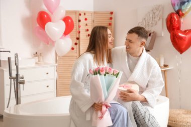 Çiçek demeti ve hediye kutuyla bornozlu güzel bir çift evde Sevgililer Günü 'nü kutluyor.