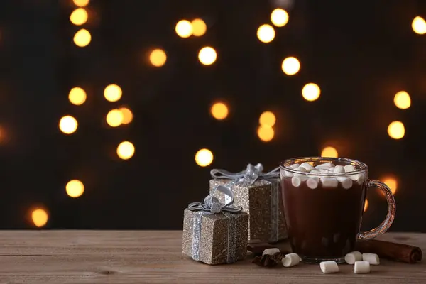 Glass cup of hot chocolate with marshmallows, different spices and gift boxes on wooden table against blurred lights