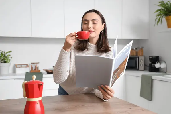 Pretty young woman with cup of espresso and geyser coffee maker reading magazine in modern kitchen