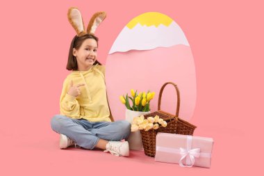 Paskalya tavşanı kulakları, sepeti, çiçekleri, hediyeleri ve pembe arka planda büyük kağıt yumurtası olan tatlı küçük bir kız.