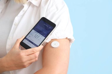 Glikoz sensörü olan şeker hastası bir kadın mavi arkaplandaki kan şekerini ölçmek için cep telefonu kullanıyor.