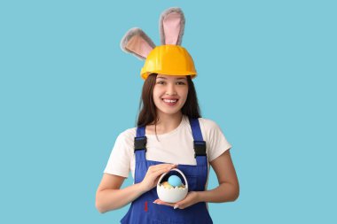 Mavi arka planda Paskalya yumurtaları olan tavşan kulaklı Asyalı kadın işçi.