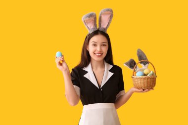 Sarı arka planda Paskalya yumurtaları olan tavşan kulaklı genç Asyalı oda hizmetçisi.