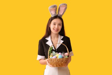 Sarı arka planda Paskalya yumurtaları olan tavşan kulaklı genç Asyalı oda hizmetçisi.