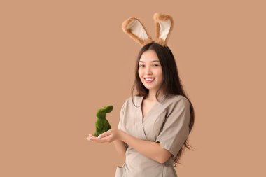 Kahverengi arka planda Paskalya tavşanı olan tavşan kulaklı Asyalı kadın masaj terapisti.