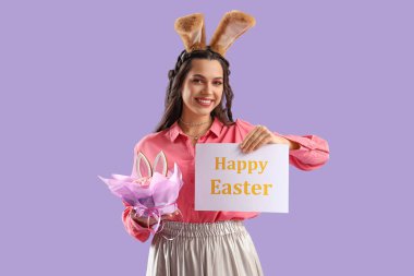 Tavşan kulaklı güzel bir kadın. Elinde kağıt var. Mutlu Paskalyalar ve leylak arkaplanlı pasta.