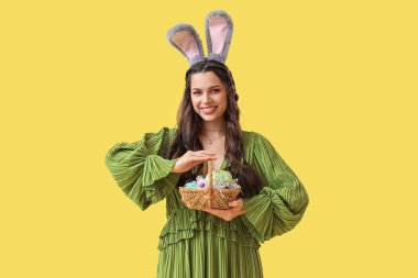 Sarı arka planda Paskalya sepeti olan tavşan kulaklı güzel bir kadın.